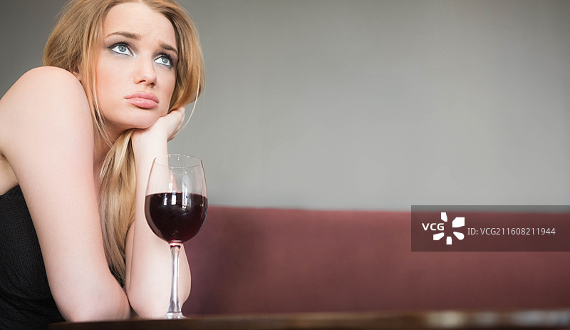 女子身穿黑色上衣，倚靠在休闲桌旁，手托着下巴向上凝视，桌上放着酒杯，旁边有留白区域。图片素材