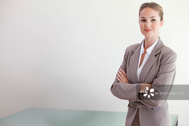 身着灰色套装的女子站在右侧墙边，双臂交叉，身旁是磨砂玻璃办公桌，留有复制空间图片素材