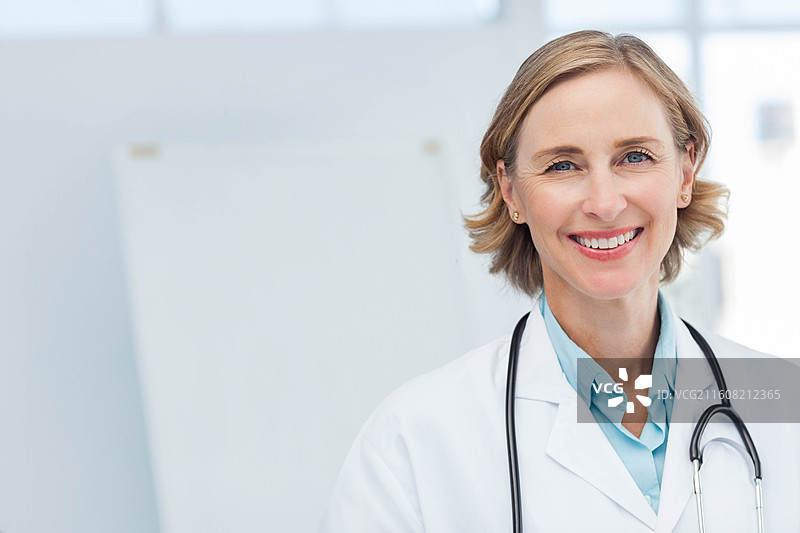 一位成熟的女医生，穿着白大褂，戴着听诊器，面带微笑看向镜头，在诊所内，有复制空间图片素材