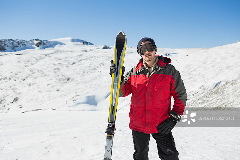 一名穿着红色夹克和护目镜的成年男性，站在雪坡上，手持黄色滑雪板，留有复制空间。图片素材