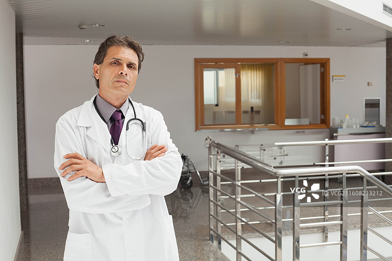 中年男性医生，身穿白大褂，佩戴听诊器，双臂交叉，站在医院里，留有复制空间图片素材