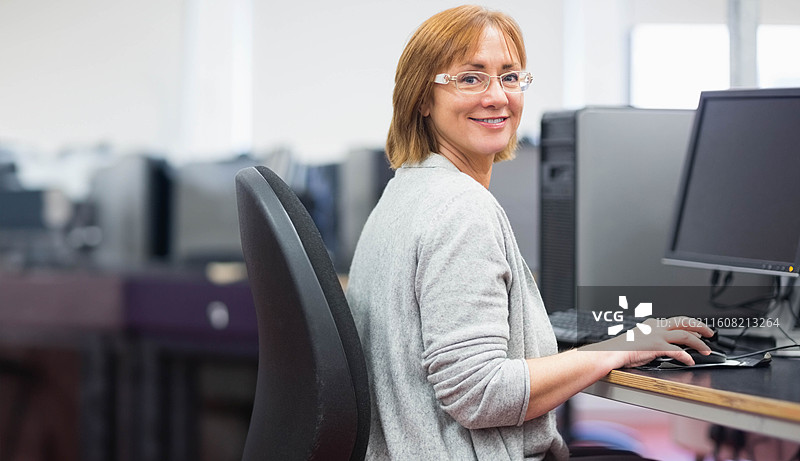 一位戴眼镜的年长女员工坐在办公桌前，使用平板显示器和鼠标图片素材