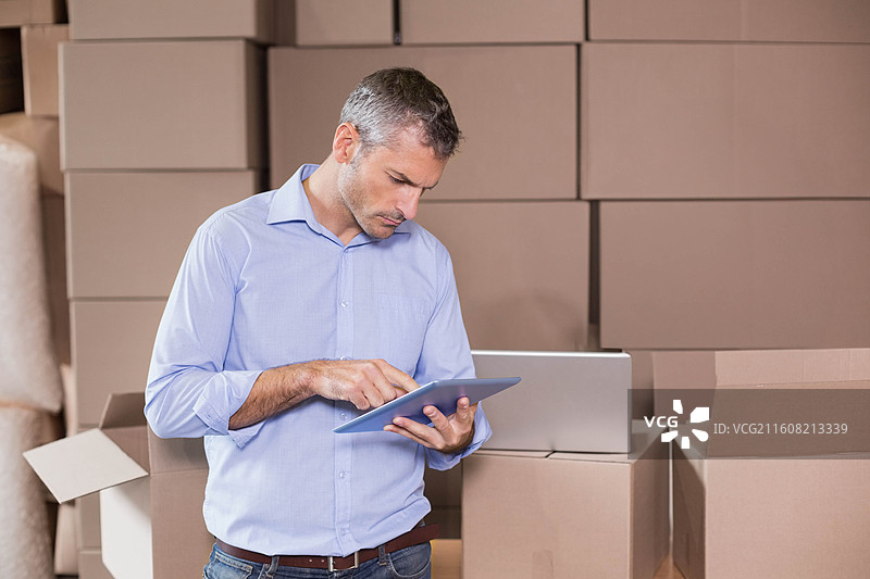 穿着蓝色衬衫和牛仔裤的中年男子在仓库的箱子中间使用平板电脑工作，旁边放着笔记本电脑图片素材