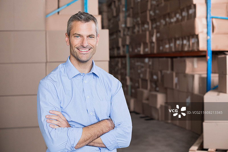 一位穿着蓝色衬衫的中年男子，双臂交叉，面带微笑地站在仓库的货架和箱子旁边。图片素材