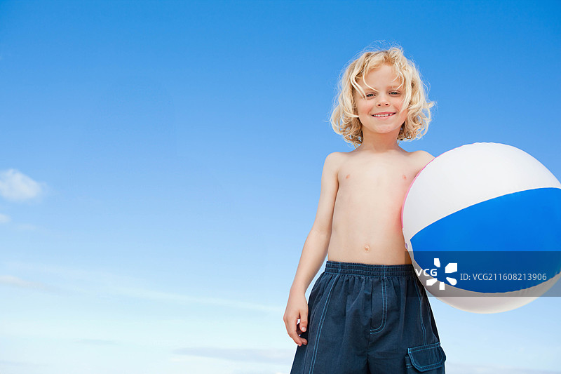 一个男孩站在海滩上，身穿深色泳裤，手持蓝白相间的沙滩球，旁边留有复制空间。图片素材