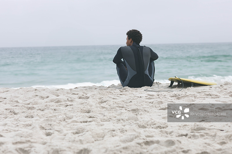 穿着深色湿衣的非洲裔美国男子坐在沙滩上，面向大海，旁边是黄色的冲浪板脚绳图片素材