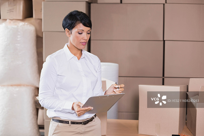 身穿白色衬衫和卡其裤的女性，手持文件夹，正在检查仓库中的箱子和气泡膜。图片素材