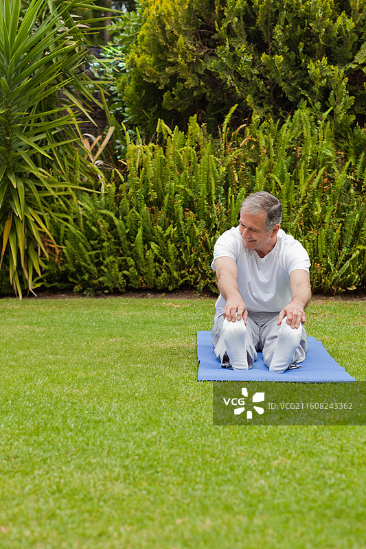 一位老年男子身穿白色T恤，在花园的蓝色瑜伽垫上做伸展运动，试图触碰脚尖，配有复制空间图片素材