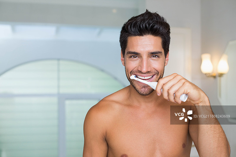 一个赤裸上身的成年男性站在现代浴室里，微笑着刷牙图片素材