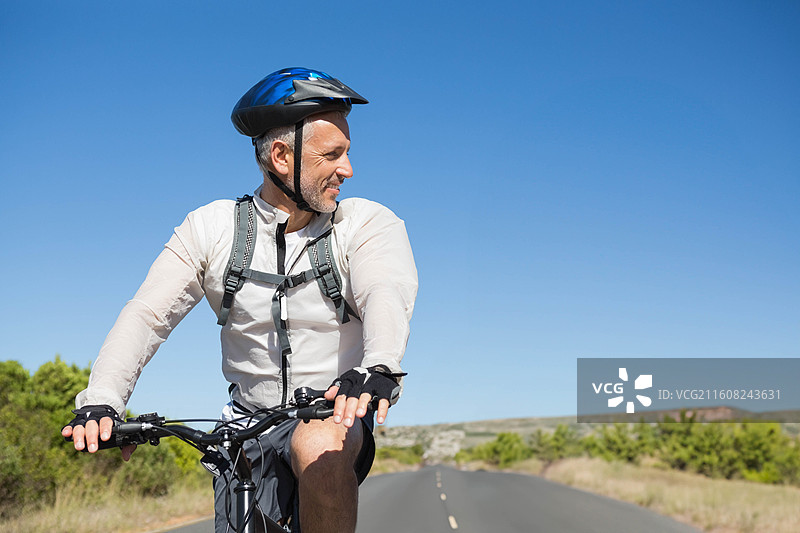 身穿蓝色头盔和背包的成熟男子在铺砌的道路上骑行公路自行车，穿过灌木丛图片素材