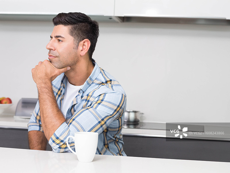 身穿格子衬衫的男人坐在白色厨房台面旁，托着下巴，手中拿着马克杯图片素材