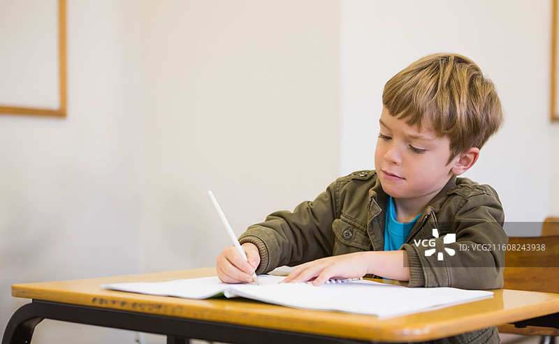 一名穿着橄榄绿夹克的男学生在学校课桌前打开笔记本写作，手里拿着一支白色钢笔图片素材