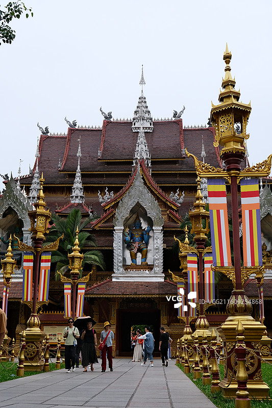西双版纳总佛寺 金色装饰立柱彩色条纹旗帜 左侧粉红花丛 远处白色尖耸建筑葱郁绿树图片素材