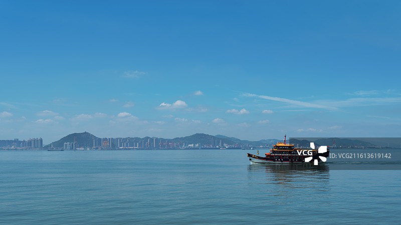 一艘装饰着黄色顶棚的游船，在厦门碧蓝的海面上航行，远处是连绵的青山与现代化的城市天际线。图片素材