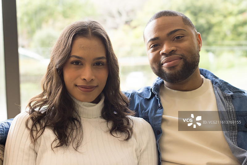 一对穿着牛仔夹克的多元化情侣在家中沙发上坐着，透过窗户欣赏花园景色图片素材