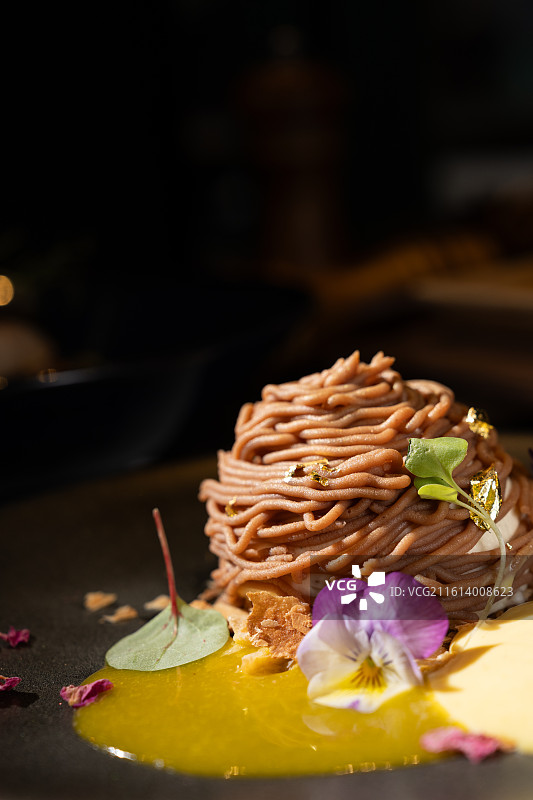 黑色餐盘上的精致蒙布朗甜点，包含栗子泥与香草慕斯，并点缀以金箔和食用花卉。图片素材