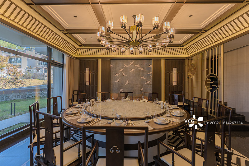 圆形餐桌放着餐具周围是多把木质椅天花板设有吊灯左侧是落地窗引入自然景观后方墙壁是飞鸟图两边是木质装饰图片素材