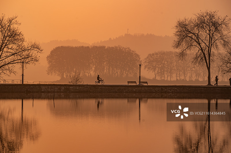武汉东湖日出晨景：橙光天际映湖岸树影水面倒影 湖畔长椅行人相伴的秋日湖景视觉画面图片素材