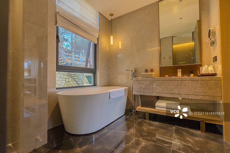 浴室内窗户旁摆有椭圆形白色浴缸旁边是大理石洗手台摆放着洗漱用品下方有放了毛巾的木质储物架墙壁挂有镜子图片素材