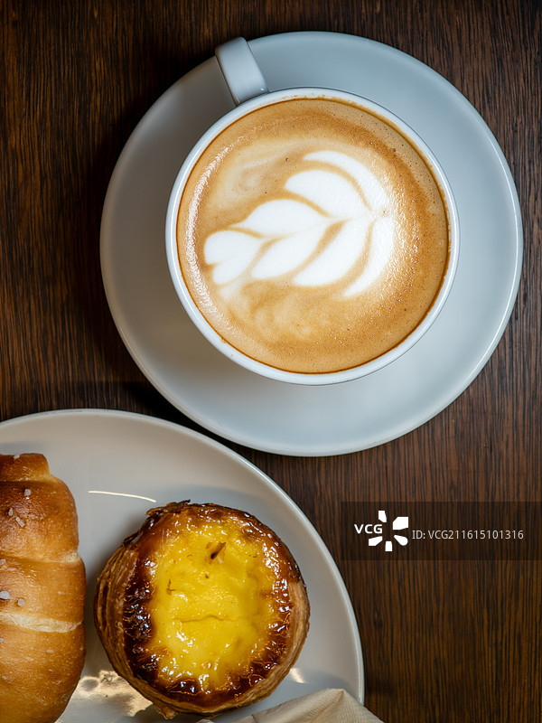 木质桌面上的拉花拿铁咖啡与蛋挞和盐面包图片素材