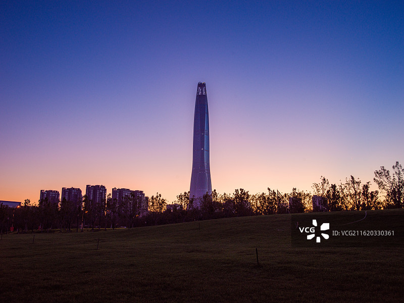 天津滨海新区高耸入云的建筑矗立在开阔的草地前，背景是渐变的绚丽天空，从柔和的粉紫到温暖的橙黄。图片素材