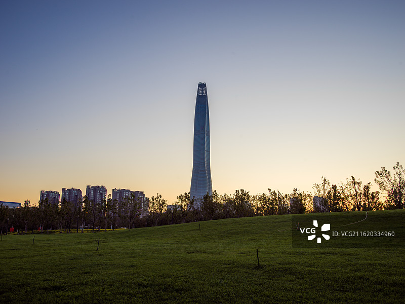 天津滨海新区高耸入云的建筑矗立在开阔的草地前，背景是渐变的绚丽天空，从柔和的粉紫到温暖的橙黄。图片素材