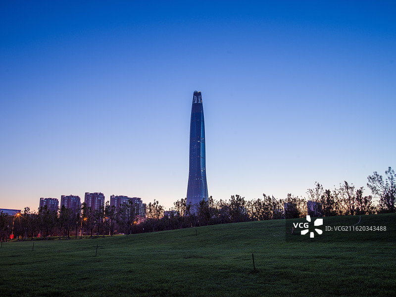天津滨海新区高耸入云的建筑矗立在开阔的草地前，背景是渐变的绚丽天空，从柔和的粉紫到温暖的橙黄。图片素材