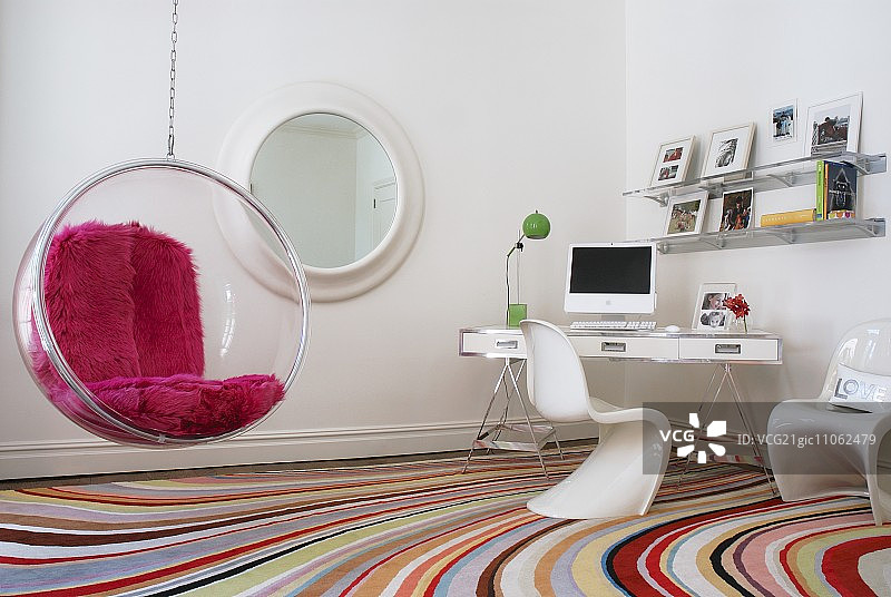 悬挂式泡泡椅，配有人造毛皮垫，位于明亮房间的角落，下方是带有弯曲条纹图案的彩色地毯图片素材