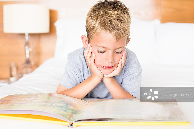 快乐的金发男孩躺在卧室的床上，在家中阅读故事书图片素材
