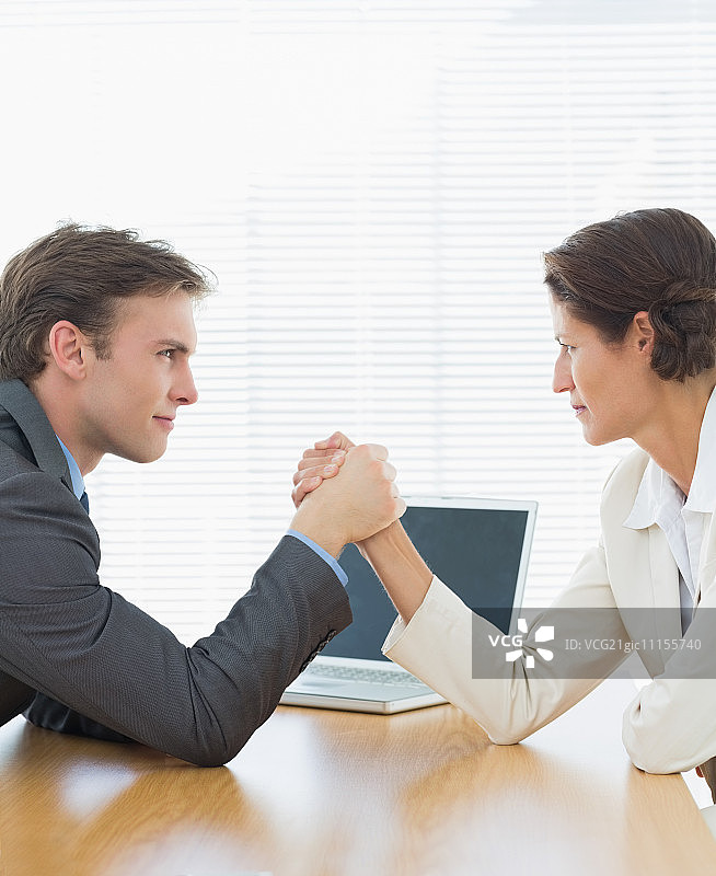 侧面观察一对表情严肃的年轻商业夫妇在办公桌上掰手腕图片素材