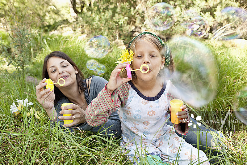 母亲和女儿（10-12岁）坐在草地上吹泡泡图片素材