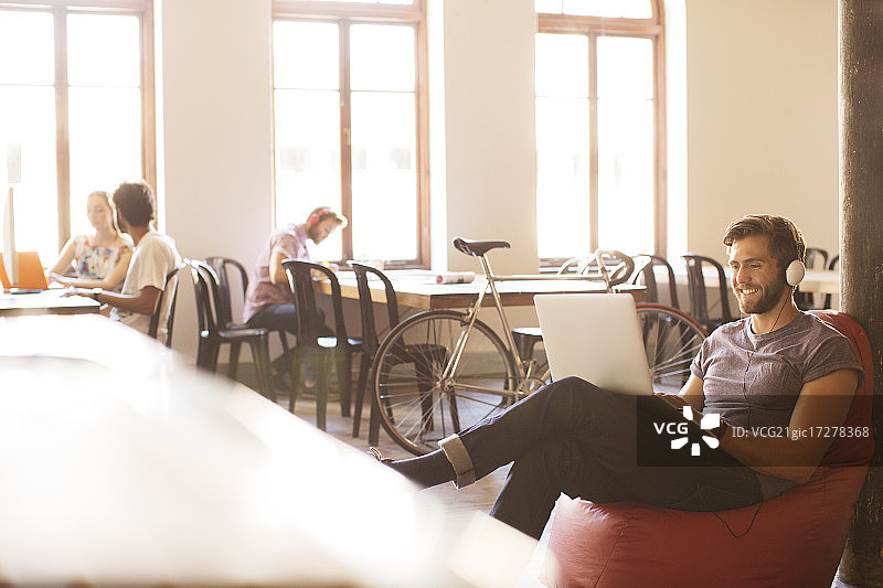 在办公室的豆袋椅上戴着耳机使用笔记本电脑工作的微笑休闲商人图片素材