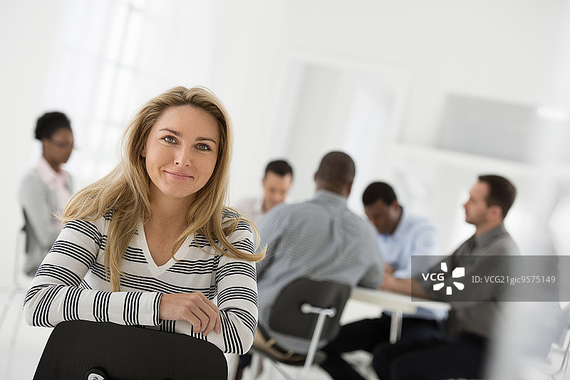 明亮洁白的建筑内部。一位身穿条纹衬衫的女子坐在椅子上，背对着一群围坐在桌旁的人。图片素材