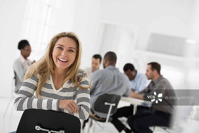 明亮洁白的建筑内部。一位身穿条纹衬衫的女子坐在椅子上，背对着一群围坐在桌旁的人。图片素材