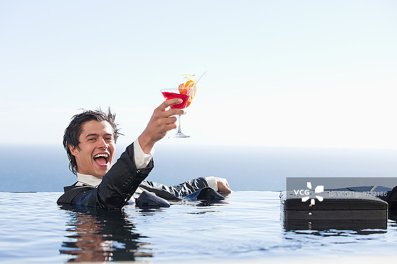 喜悦的商人穿着西装在游泳池里放松，手持鸡尾酒图片素材