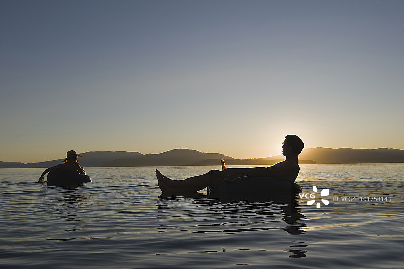 年轻情侣湖边日落泳圈漂浮，享受冷饮图片素材