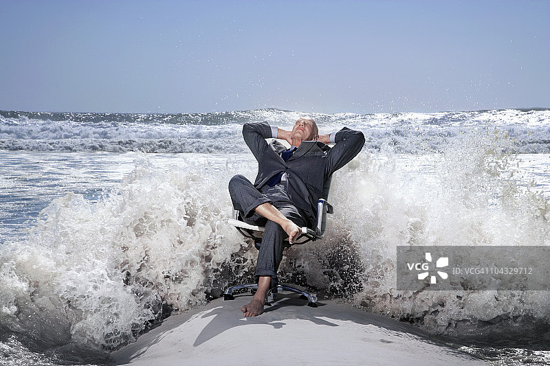 年长的商人在沙滩上的办公椅上，被海浪拍打图片素材