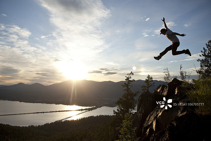 日落时分，年轻男人在俯瞰湖泊的悬崖边翱翔图片素材