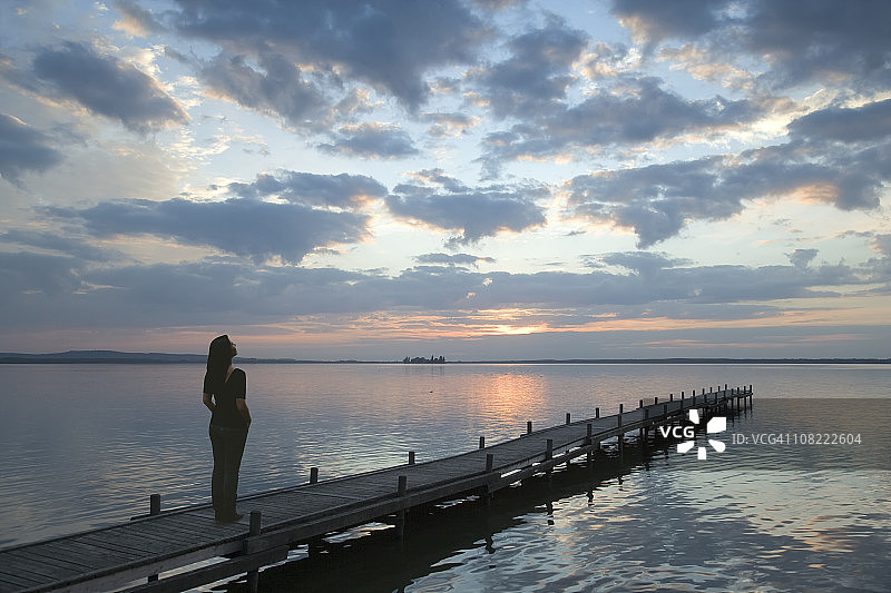 站在湖边码头，欣赏日落云景的年轻女人图片素材