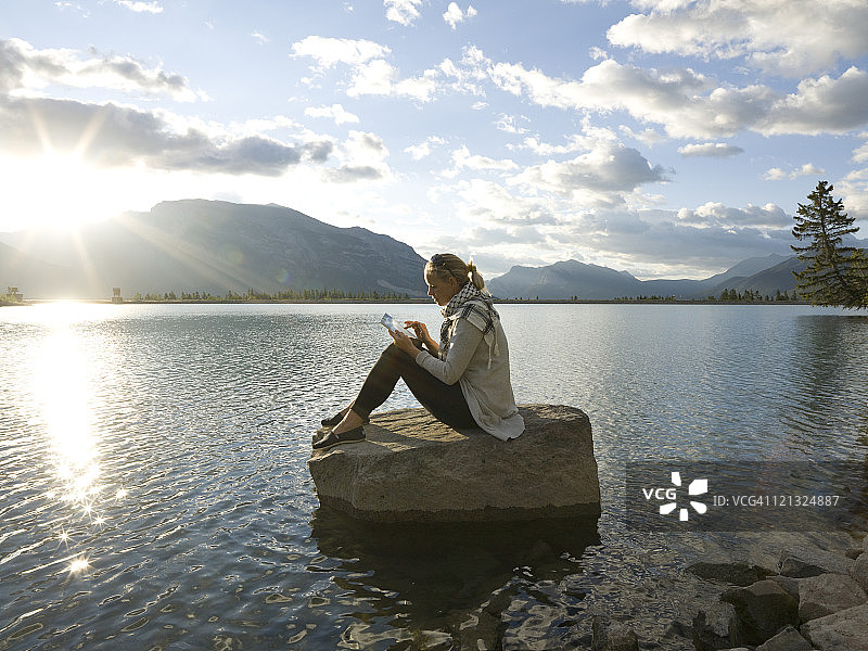 日出时分，女人坐在岩石岛上，使用平板电脑工作图片素材
