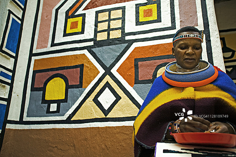 恩德贝勒族妇女身着已婚妇女的传统服装。背景是恩德贝勒村庄的传统几何墙绘，南非约翰内斯堡附近的莱塞迪文化村图片素材