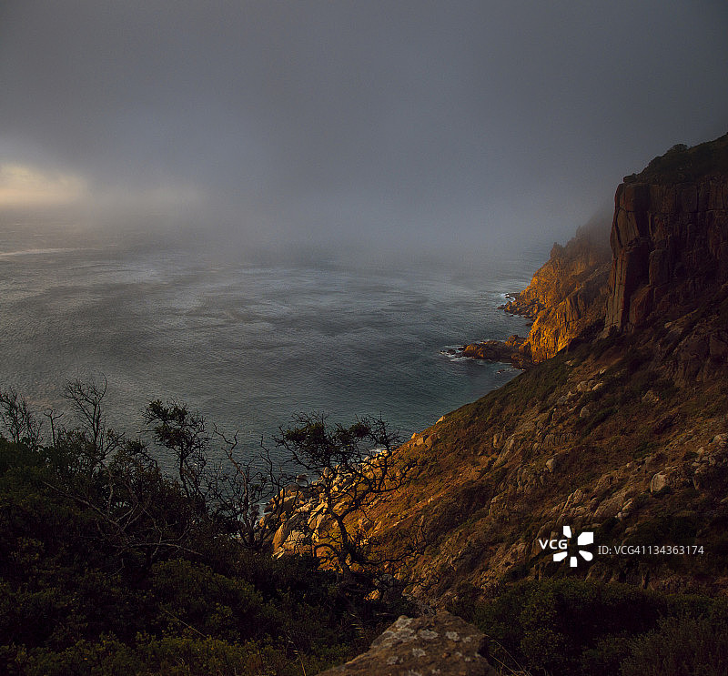 南非西开普省开普敦查普曼峰，在低矮的阴暗薄雾云层下，夕阳照耀着海洋，岩石海岸线前斜坡上生长着稀疏的树木，呈现出明亮的橙红色图片素材