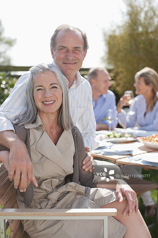 在阳光明媚的花园里与朋友在桌旁享受午餐的微笑老年夫妇的肖像图片素材