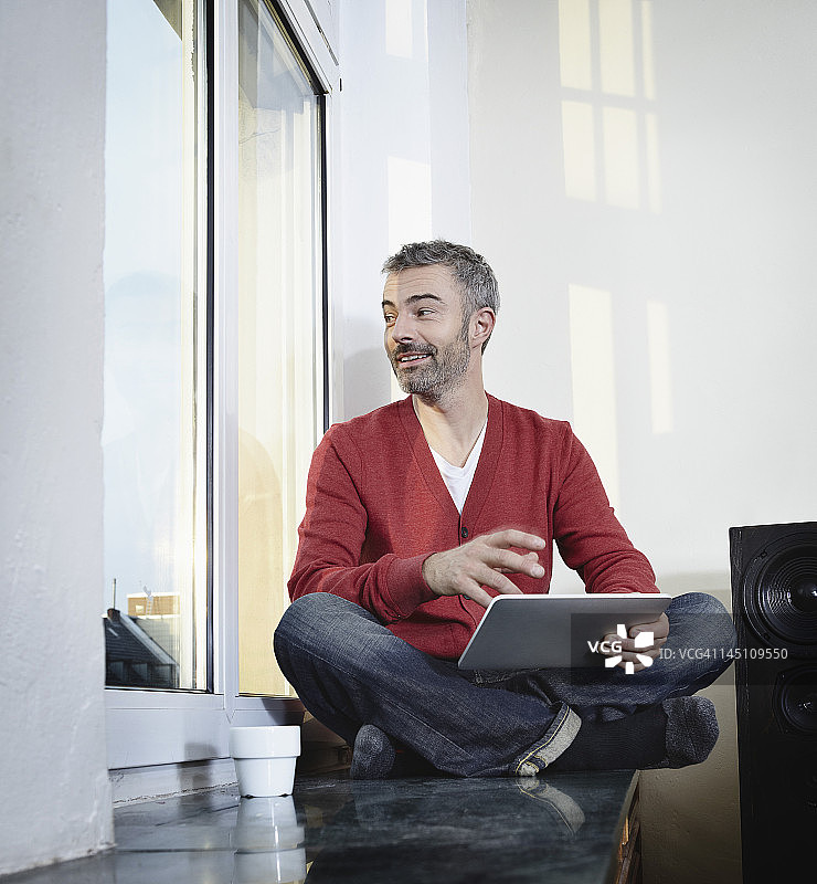 德国科隆：中年男子坐在窗边使用平板电脑，微笑图片素材