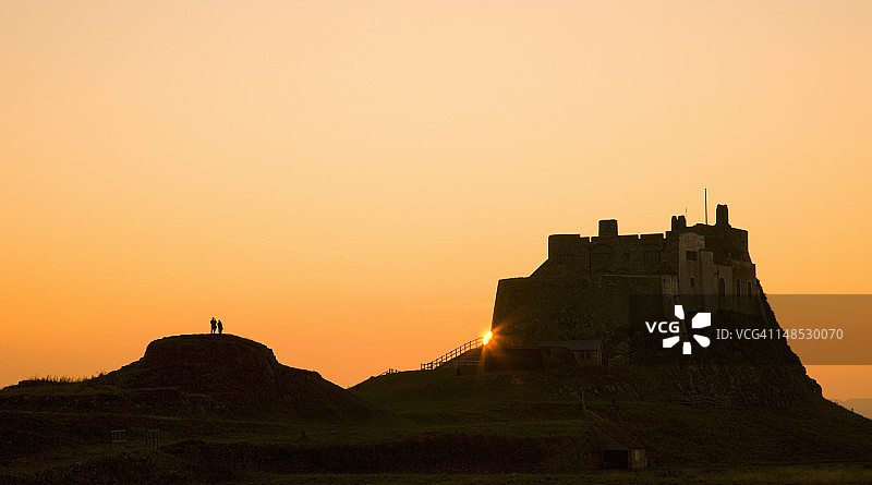 林迪斯法恩城堡后的日落与岩石上的游客剪影图片素材