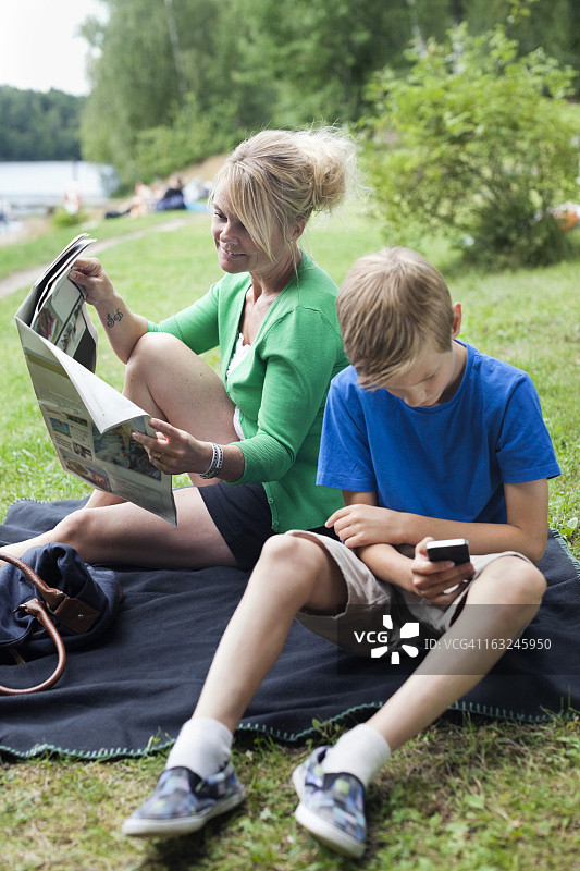 在公园里，中年妇女和使用手机的儿子一起看报纸图片素材