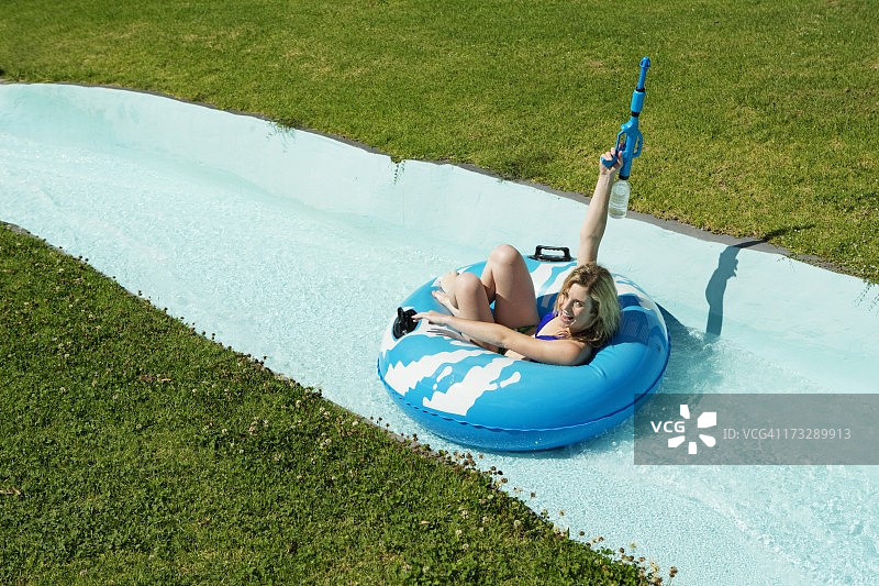 年轻女子坐在游泳圈上，手持水枪滑下水滑梯图片素材
