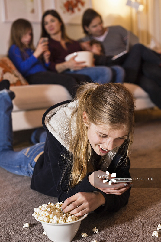 快乐的女孩在地板上吃着爆米花，家人坐在沙发上看手机图片素材