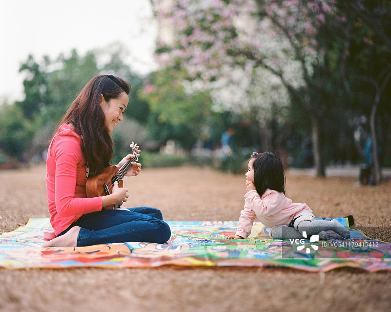年轻妈妈在草坪上为幼儿弹奏尤克里里图片素材