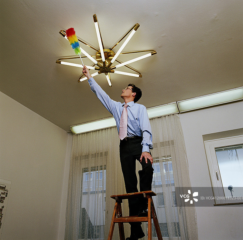 商人站在梯子上用鸡毛掸子清洁灯具图片素材
